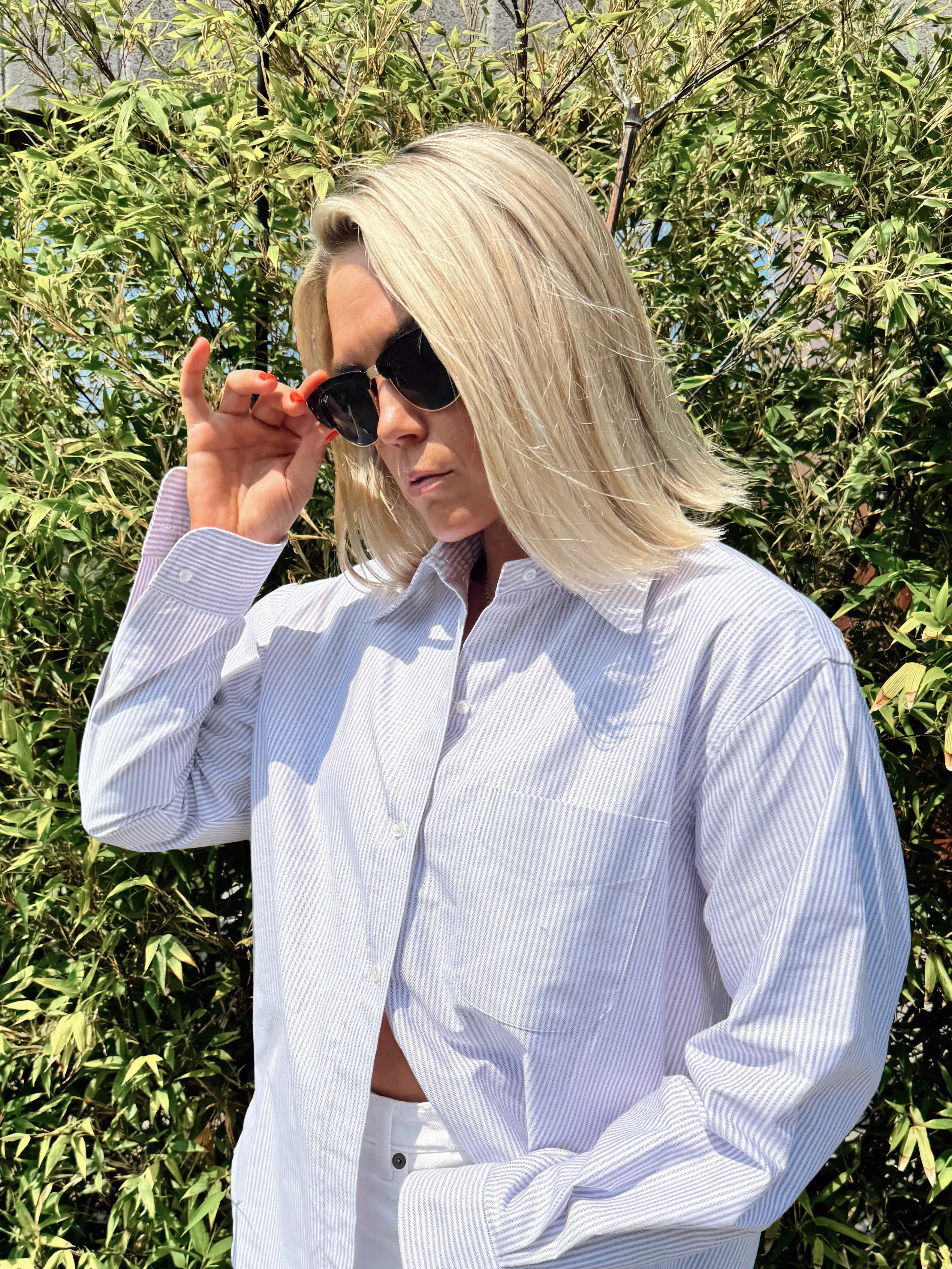 Person wearing a light purple striped shirt with sunglasses, standing against a green leafy background