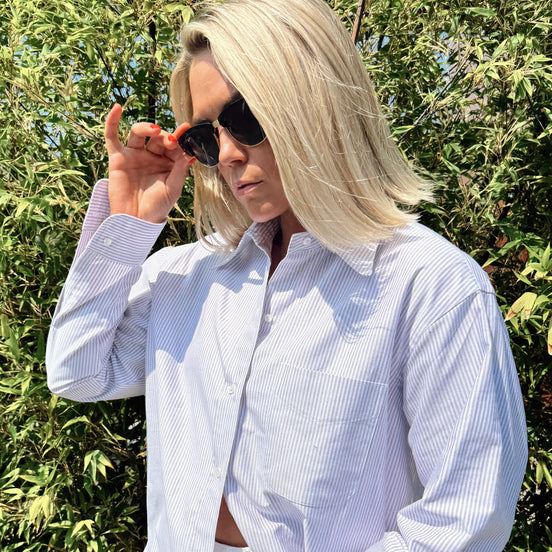 Person wearing a light purple striped shirt with sunglasses, standing against a green leafy background